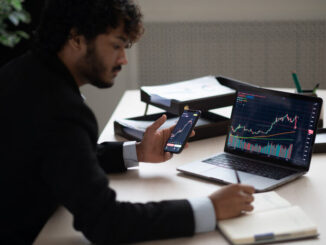 Smart focused Indian broker investor, stock trader using smartphone and laptop to analyze and invert in the financial market of cryptocurrencies, buy or sell crypto coins, planning strategy